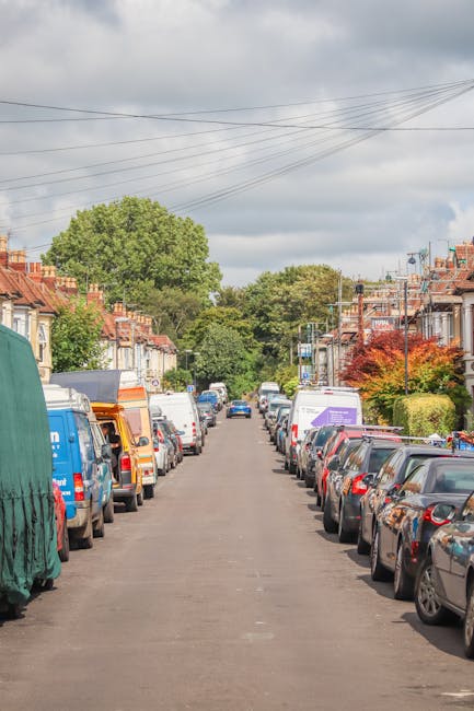 A residential street scene during daytime shows a narrow, straight road lined with parked cars on both sides, including sedans, hatchbacks, and vans. At the foreground, a large white van is partially visible, with its side door open, suggesting loading or unloading activities. The van is parked close to the curb on the right side, near a house with a front garden. Several cardboard boxes and wrapped furniture pieces are visible on the pavement near the van, indicating a home relocation process. The road appears to be in a quiet neighbourhood with terraced houses featuring brick facades, small front gardens, and bay windows, some with colorful foliage suggesting seasonal changes. Overhead, electrical wires run parallel to the street, and in the background, greenery and large trees are visible, with a cloudy sky overhead. The scene reflects a typical house removal or moving operation managed by Man with Van The Hyde, highlighting the logistics involved in furniture transport and packing during a move.