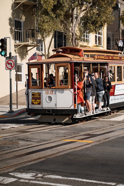 A vintage-style tram painted in cream and brown, with open windows and a sign displaying 'Powell & Hyde Sts,' is travelling along tram tracks on a city street. Several passengers, including a woman in a red coat and a man in a dark jacket, are standing on the tram's open platform at the rear, holding onto the railing as they prepare to disembark or board. The tram is surrounded by urban buildings with balconies, trees, and a street light, while pedestrians wait to cross the road. This scene captures an urban transportation method often used for sightseeing or local travel, illustrating a typical day in a city with historical transit options, relevant to house removals and moving services provided by Man with Van The Hyde, supporting home relocation and furniture transport processes.