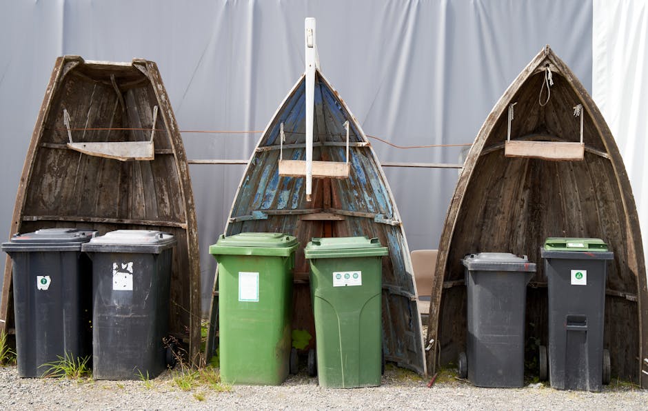 Three wooden boat hulls positioned upright outdoors, used as makeshift storage or waste containment, with the middle boat painted blue and the others left natural wood, situated behind a row of four wheelie bins in black and green, each labeled for rubbish or recycling. The bins are placed on a gravel surface, in front of the boat hulls, near a neutral-colored fabric backdrop. Behind the bins, a white boundary line or clothesline is visible, stretching across the scene, which may be related to waste organization or outdoor storage. The overall scene depicts waste collection and disposal components, with the boat hulls possibly repurposed or waiting for removal, reflecting elements relevant to house removals, packing, and moving logistics, as managed by Man with Van The Hyde.