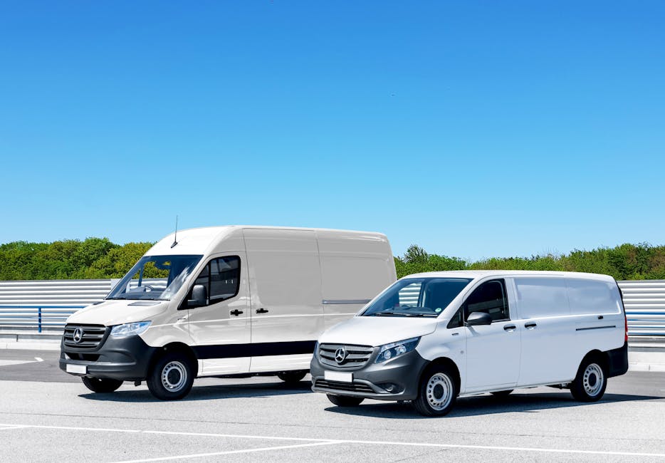 A small white commercial van parked on the side of a street with a smooth, paved surface and white road markings. The van is positioned perpendicular to the sidewalk, with the driver visible inside, p