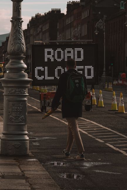 A person with a dark backpack, dressed in a black jacket and beige trousers, is walking across a city street toward a large electronic road closure sign that displays the words 'ROAD CLOSED' in bright white lights. The sign is situated on the pavement, behind a row of orange and white traffic cones, indicating a temporary traffic restriction likely due to roadworks or events. To the left, there is an ornate street lamp with intricate detailing on its base, and the surrounding environment includes red-brick residential buildings with multiple windows visible in the background. The scene is set in daylight with overcast skies, and the street appears to be part of a busy urban area, possibly near a house removal or relocation service, as indicated by the context of Man with Van The Hyde. This image captures a moment during a relocation project where street closures impact access, relevant to logistics and transportation for home or furniture moves.