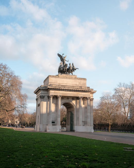 Photograph of a large stone archway on a paved pathway, with a decorative statue of a chariot drawn by horses and a rider on top, situated in a park-like setting with leafless trees on either side. The surrounding area features green grass, and in the background, there are groups of people walking along the pathway. The sky is partly cloudy with patches of blue, and the scene appears to be during daytime. This setting may be relevant to home relocation or moving services offered by Man with Van The Hyde, illustrating an outdoor environment where furniture or boxes could be temporarily arranged during a moving process.