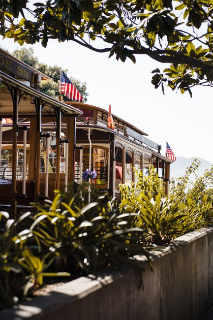 A vintage-style tram with wooden paneling and decorative metal railings is partially visible in the image, parked alongside a stone wall that is topped with lush green foliage and potted plants. The tram is adorned with small American flags attached to its exterior, and its interior, visible through the windows, contains various items including blankets and packing materials, suggesting preparation for a home relocation or moving process. The surrounding environment includes natural daylight and overhanging tree branches, creating a pleasant outdoor setting. The scene reflects the logistical aspects of furniture transport and packing involved in house removals, with the tram positioned on a paved area adjacent to a garden or landscaped space. This setting exemplifies the careful handling and transport elements associated with professional removals services, such as those offered by Man with Van The Hyde, specializing in emergency same-day moves in The Hyde area.