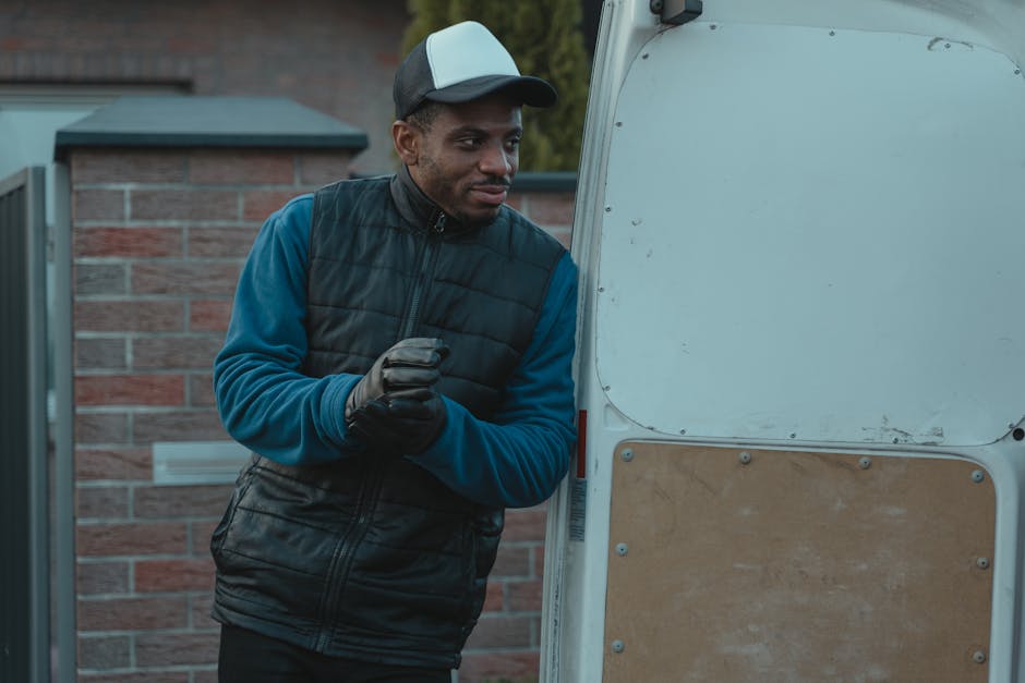 A man is standing outside near a moving van, dressed in a black and grey cap, a navy blue long-sleeve shirt, and a black padded vest. He is wearing gloves and appears to be in the process of preparing for a home relocation, with a serious expression on his face. The rear door of the van is open, revealing a partially visible interior used for furniture transport and packing materials such as cardboard boxes and plastic wrap visible on the ground or nearby. The van is parked adjacent to a brick wall of a residential property, with some greenery in the background. The scene captures the loading process common to house removals, involving careful handling of items and equipment like moving blankets and trolleys, typical of services provided by Man with Van The Hyde, supporting smooth relocation and luggage transport. Natural lighting indicates it may be early morning or late afternoon, with a focus on the preparation steps involved in a professional delivery and moving service.