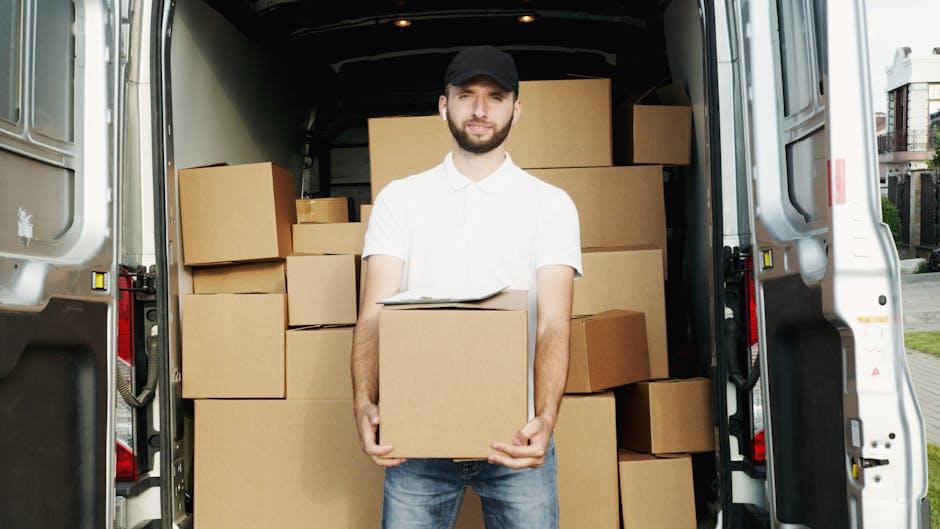 A young man with a beard, wearing a white polo shirt, black cap, and jeans, stands inside the open rear of a large moving van, holding a cardboard box. The van’s interior is filled with neatly stacked cardboard boxes of various sizes, some wrapped in plastic or fabric, ready for home relocation. The loading area beside the van shows pavement and a residential street with houses and greenery in the background, indicating an exterior setting. The scene captures the process of furniture transport and packing during a house removal, with the man preparing items for unloading or loading. The professional appearance and organized packing materials underscore services provided by Man with Van The Hyde, aligned with the house removals category on the website.