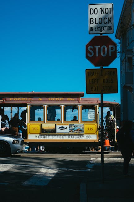A vintage yellow and white trolley bus with open sides is stopped at a street intersection near Hyde Town Hall, serving the Powell and Hyde Sts routes. Several passengers are visible seated inside, some standing, with dark silhouettes against the glass. The bus is positioned behind a metal barrier and is surrounded by moving and parked vehicles, including a silver car partially visible on the left. Above, multiple traffic signs are mounted on poles, including a 'Do Not Block Intersection' sign, a red STOP sign, and a yellow warning sign indicating left-door traffic. The background features a blue sky and parts of buildings, suggesting a city street scene during daylight hours. The environment is consistent with urban transportation areas where home relocation and furniture transport services by companies like Man with Van The Hyde are often required for local moving projects connected to house removals near Hyde Town Hall.