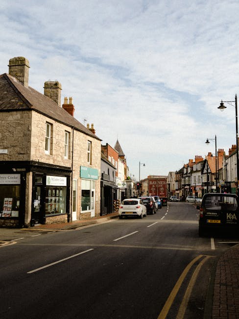 A street scene in a small town featuring a row of mixed-use buildings with stone and brick facades, some with shopfronts and signage, under a partly cloudy sky. Parked cars line both sides of the narrow road, with a white vehicle positioned near the curb. On the right, a man from Man with Van The Hyde is seen loading or unloading large cardboard boxes, wrapped in plastic and positioned on dollies or flat carts, indicating a home relocation or furniture transport process. The area is illuminated by vintage-style street lamps, and the scene captures an active moving operation as part of a local house removal service near Hyde Town Hall, supporting efficient packing and loading during the logistics of house removals.