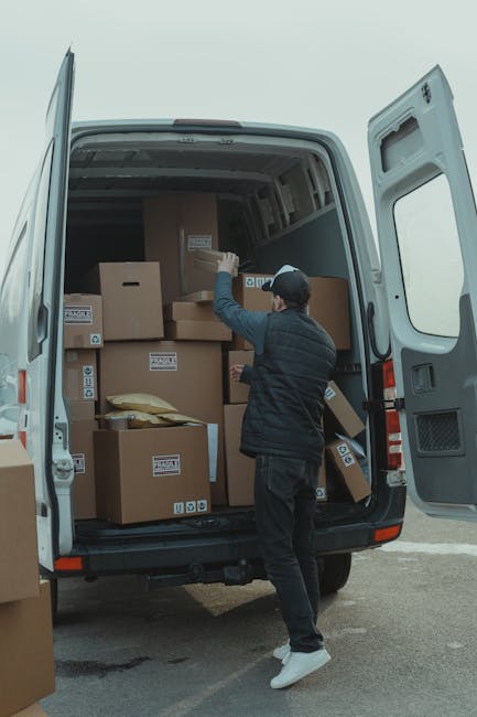 A young man with a beard and dark hair, wearing a red baseball cap, a brown and beige jacket over a checked shirt, and dark jeans, is sitting on the edge of an open van's loading area. Behind him, the