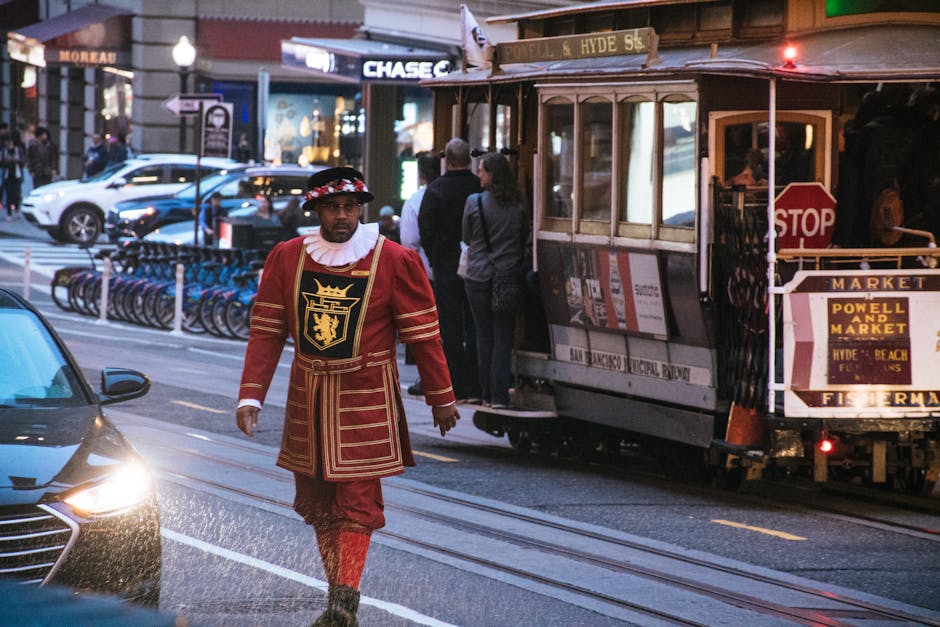 A man dressed in traditional Tudor-style costume with a red and gold embroidered tunic, matching tights, and a hat with a red and white floral band, walking along a street in an urban area during dusk. Behind him is a classic tramcar with a wooden exterior, open sides, and a stop sign, indicating a tram stop nearby. The tram displays signage for Powell and Market Street, and is parked on railway tracks running along the road. To the left, a modern black vehicle is partially visible with its headlights turned on, and several motorcycles are parked in front of a building with illuminated signage that reads 'Chase' and other shops, suggesting a busy city environment. Pedestrians, some carrying bags, wait at the tram stop beyond the man, and streetlights cast a warm glow on the scene. The environment suggests an active city street suitable for home relocation and furniture transport involving careful handling of packed boxes and furniture moving logistics, as seen through the presence of the tram and urban infrastructure.