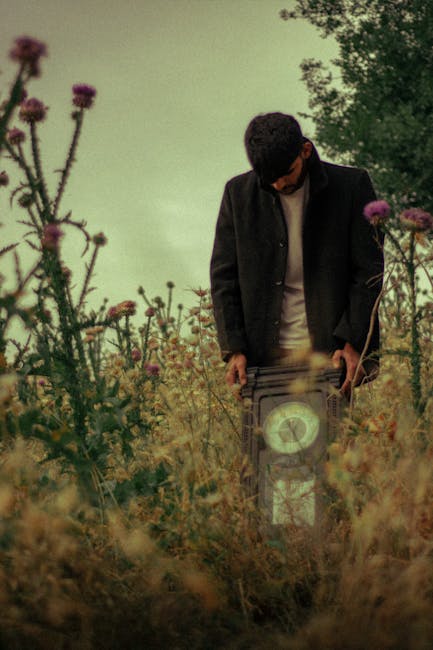 A man dressed in dark clothing and wearing sunglasses is standing outdoors among tall wildflowers and grasses. He is holding a vintage-style alarm clock in his right hand, which is positioned near his thigh. The scene features natural lighting with a cloudy sky overhead, and the man appears to be engaged in a symbolic or artistic act related to time. The surrounding environment suggests a rural or garden setting. This image reflects themes associated with home relocation and moving logistics, with a focus on the passage of time. The subdued colors and thoughtful composition align well with professional removals services, such as those offered by Man with Van The Hyde, as they relate to packing, transporting, and managing belongings during house moves.
