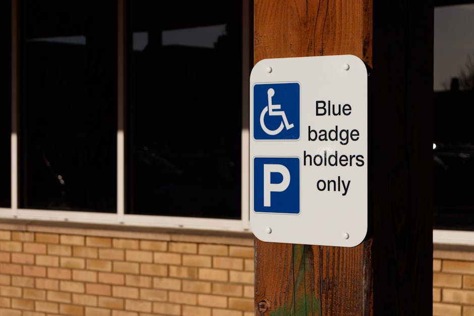 A close-up image of a rectangular white parking sign mounted on a brown wooden post outdoors near a brick building with large black-tinted windows. The sign features a blue disability symbol at the top, indicating accessibility, followed by the text 'Blue badge holders only' in black lettering. Below this, there is a blue square with a white 'P,' denoting parking. The setting appears to be in daylight, and the sign is positioned at eye level, likely to guide disabled drivers or residents in a residential area. This image is relevant to house removals and relocation services provided by Man with Van The Hyde, illustrating parking regulations that may be encountered during home relocation or furniture transport in The Hyde, as outlined in the webpage about Tameside Council parking permits.
