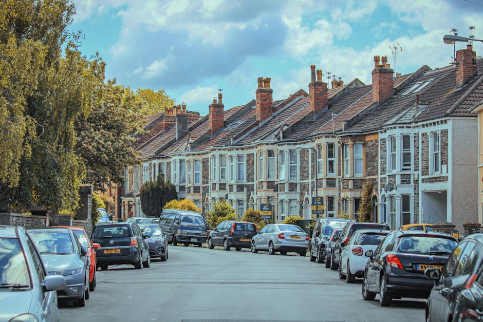 A white moving van parked on a narrow, sloped street surrounded by multi-storey residential buildings in an urban area during daylight. The van is positioned close to the curb with its front facing uphill, and partially blocking the road. Next to the van, there are a few scattered cardboard boxes and wrapped furniture items suggesting an ongoing home relocation process. The surrounding buildings are constructed with brick and concrete, with some featuring external fire escapes and multiple windows. A black trash bin is visible on the pavement near the van, and a red car is parked further down the street. Shadows from the buildings cast across the road, and a partly cloudy sky overhead. This scene illustrates a typical urban furniture transport and loading process suitable for house removals, with [COMPANY_NAME] likely involved in the logistics of packing, loading, and transporting belongings in the context of moving in and out of properties in The Hyde area.
