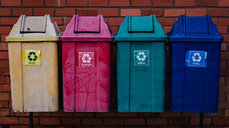 Four large recycling bins placed side by side against a red brick wall, each with a different colour and label indicating the type of waste: the first is yellow with a label for 'METAL,' the second is pink with 'PLÁSTICO,' the third is teal with 'VIDRO,' and the fourth is blue with 'PAPEL.' The bins are positioned outdoors on a paved area, and their lids are closed. The yellow bin shows noticeable dirt and some graffiti on its surface. This scene relates to waste disposal guidelines in The Hyde, as part of the house removal and packing process, ensuring proper sorting of recyclables during a home relocation, with Man with Van The Hyde providing professional services for such activities.
