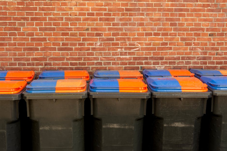 Several large black wheelie bins with orange and blue lids are lined up along a brick wall, which serves as the background. The bins appear to be positioned outdoors on a pavement or sidewalk, and are used for waste disposal. The lids are closed, and the bins are uniformly arranged in a row, indicating an organized disposal area. This setting is relevant to house removals and moving services provided by Man with Van The Hyde, as proper waste management and disposal are important aspects of home relocation and moving logistics, especially when adhering to local regulations like Waste disposal rules in The Hyde, Tameside Council guidance.